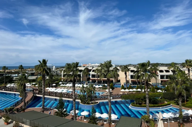 Widok na luksusowy kompleks hotelowy z basenami otoczonymi palmami, z błękitnym niebem w tle. Widok na luksusowy kompleks hotelowy w Riwierze Tureckiej, z basenami otoczonymi palmami, z błękitnym niebem w tle.