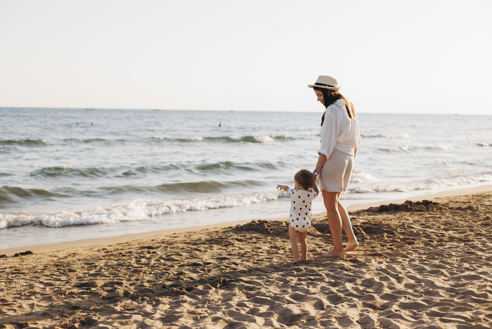 Wakacje z niemowlakiem – czy warto i jak się przygotować? 8 Mama z córką obserwują fale na pięknej plaży.
